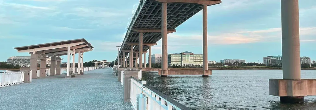 View along the paved walkway of Mount Pleasant Pier under the Arthur Ravenel Jr. Bridge, with concrete support columns, white railings, covered shade structures, and distant buildings across the water under a blue sky with light clouds.
