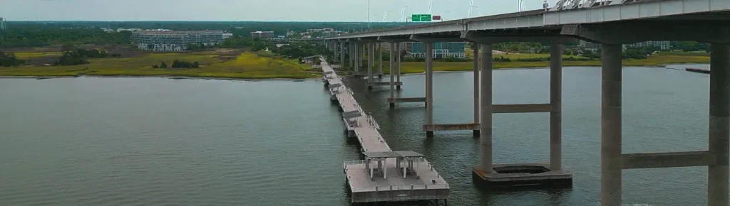 Elevated overview of the long Mount Pleasant Pier extending into the water alongside the Arthur Ravenel Jr. Bridge, with multiple shade pavilions on the pier, surrounding marsh and buildings on the left shoreline under a cloudy sky.