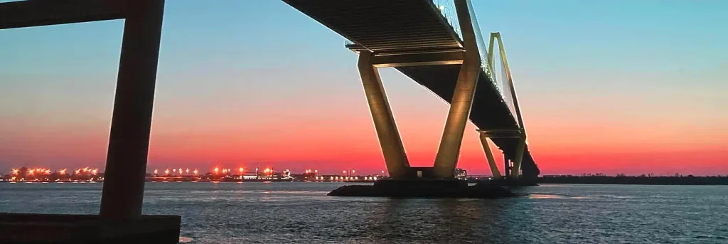 Sunset silhouette of the cable-stayed Arthur Ravenel Jr. Bridge from the water/pier level, with glowing orange and red sky, illuminated bridge lights, industrial lights on the distant shore, and a large concrete pillar in the left foreground.