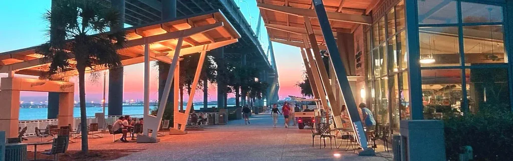 Evening scene at the base of the pier/park with warm lighting under wooden shade structures and palm trees, outdoor seating and people dining or walking, the illuminated Arthur Ravenel Jr. Bridge in the background, and a glass-fronted building (likely the River Watch Café area) on the right.
