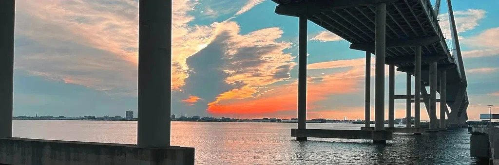Dramatic sunset view from under the bridge pillars at Mount Pleasant Pier, with orange and pink clouds reflecting on the calm water, the bridge structure extending to the right, and distant city lights/skyline on the horizon.