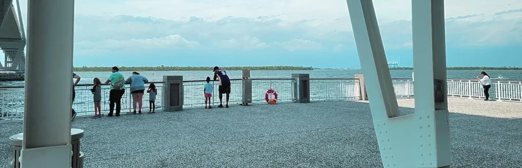People (adults and children) leaning on the white railing of Mount Pleasant Pier looking out at the harbor, with large white bridge support structures framing the scene, a pink life ring, and distant shoreline.