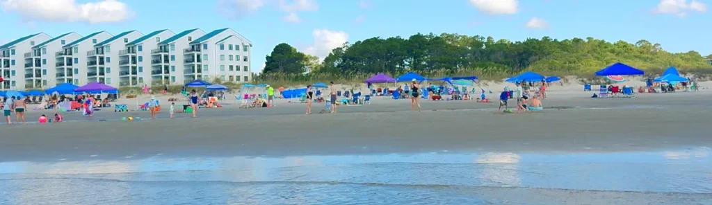 Busy beach scene at Myrtle Beach with people, colorful umbrellas and tents, multi-story condo buildings, and dunes under a blue sky.