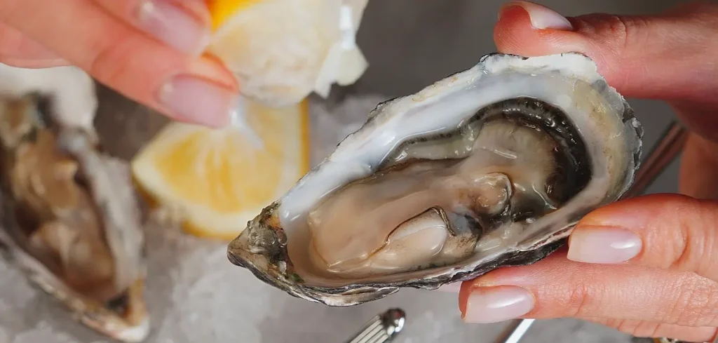 Hand squeezing fresh lemon over a raw oyster on the half shell