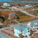 Aerial view of elevated coastal homes on stilts along Oak Island, North Carolina, with sandy lots, marshes, and the Intracoastal Waterway in the background.