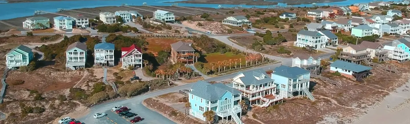 Aerial view of elevated coastal homes on stilts along Oak Island, North Carolina, with sandy lots, marshes, and the Intracoastal Waterway in the background.