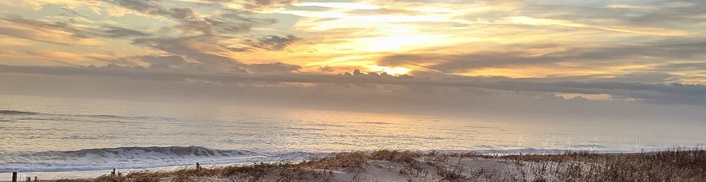 Soft golden sunrise or sunset over the Atlantic Ocean viewed from the dunes, with gentle waves, sea oats in the foreground, and layered clouds reflecting warm light.