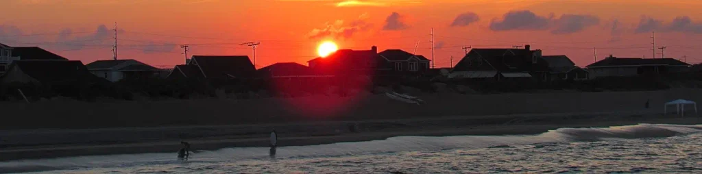 Dramatic orange-red sunset over beach cottages and dunes on the Outer Banks, with silhouetted houses, a few people on the sand, and gentle waves in the foreground.