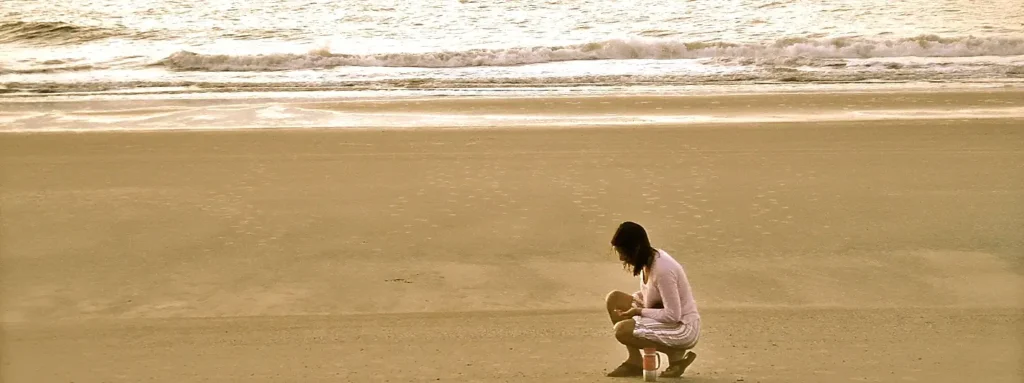 A woman crouches to collect shells on the shoreline at golden hour