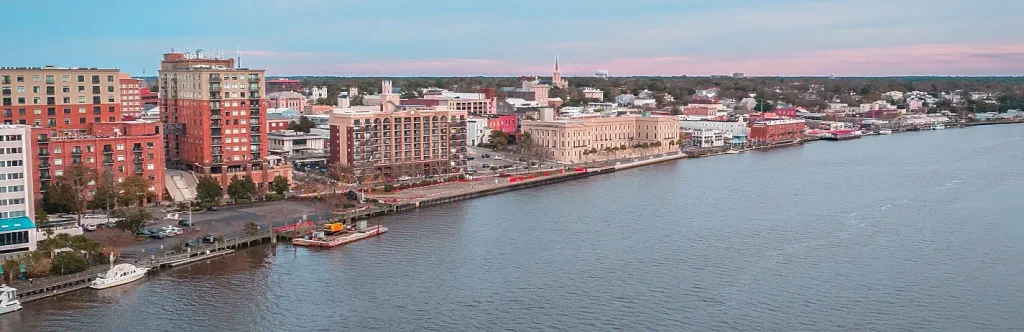 Aerial view of Wilmington, North Carolina’s waterfront skyline with multi-story brick and modern buildings, a riverwalk, boats, and the Cape Fear River under a soft sunset sky.