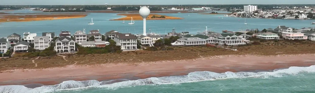 Aerial view of Wrightsville Beach, North Carolina, showing beachfront homes, a marina, a tall white water tower, tidal marshes, and the Atlantic Ocean with sailboats.