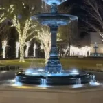 A classical multi-tiered fountain lit in blue at night in a Charleston plaza or park, surrounded by bare trees wrapped in warm white string lights, with outdoor seating and a few people walking nearby.