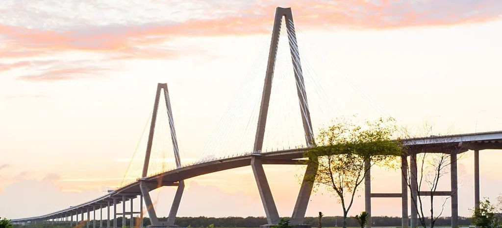 The cable-stayed Arthur Ravenel Jr. Bridge spanning the Cooper River in Charleston, SC, viewed at sunset with warm orange and pink skies, its tall A-frame towers and radiating cables silhouetted against the light.