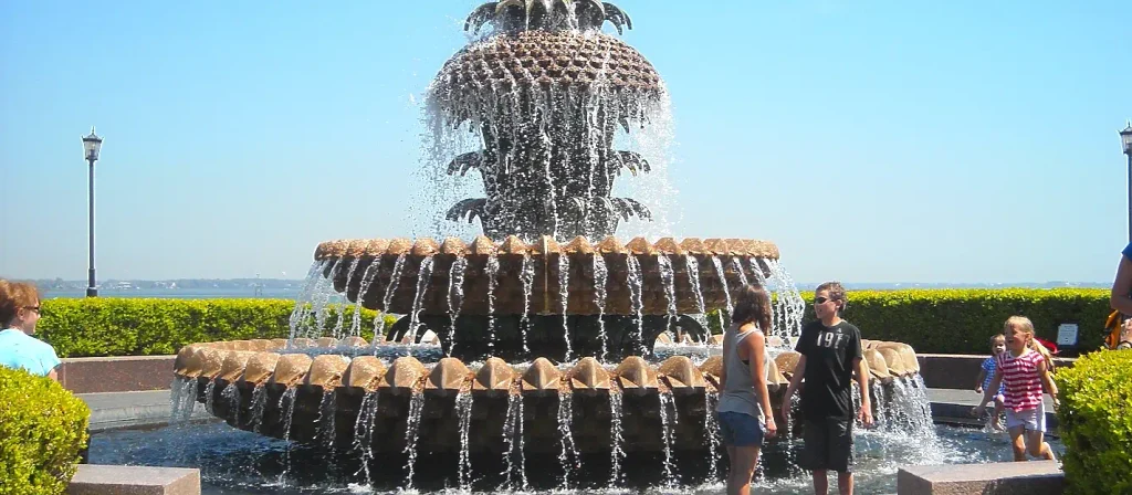The iconic multi-tiered Pineapple Fountain in Waterfront Park, Charleston, SC, with water cascading over its pineapple-shaped top and shell-like basins; people stand nearby under a clear blue sky with the harbor in the background.