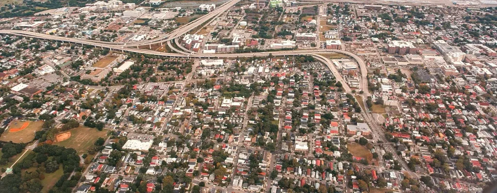 Aerial view of a dense residential neighborhood in Charleston, South Carolina, with tree-lined streets, colorful rooftops, a baseball field in the lower left, and elevated highways and interchanges cutting through the urban landscape.