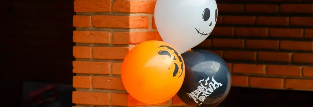 Orange jack-o’-lantern balloon, white ghost balloon, and black “Happy Halloween” balloon tied to a red-brick column, ready for a coastal Carolina Halloween porch or festival entrance.

