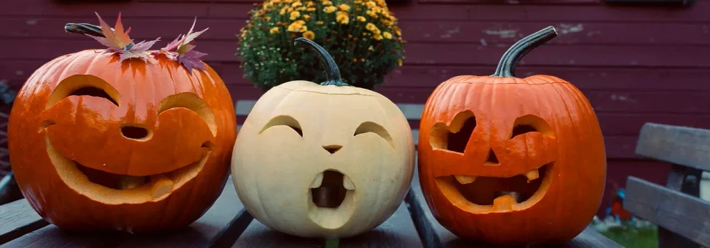 Trio of carved pumpkins on a wooden table — a smiling orange one topped with autumn leaves, a pale open-mouthed jack-o’-lantern, and a classic toothy orange pumpkin — perfect for a Carolina coast fall display or pumpkin-carving party.

