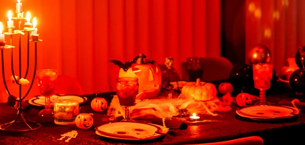 Dimly lit red-hued Halloween table set with a glowing jack-o’-lantern, candelabra, face-printed plates, spiderweb accents, and spooky glassware — a festive coastal Carolina dinner party ready for guests.

