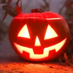 Illuminated jack-o’-lantern sitting on sandy ground amid dry coastal plants and soft fog, evoking a spooky evening walk or yard display near the Carolina shore.