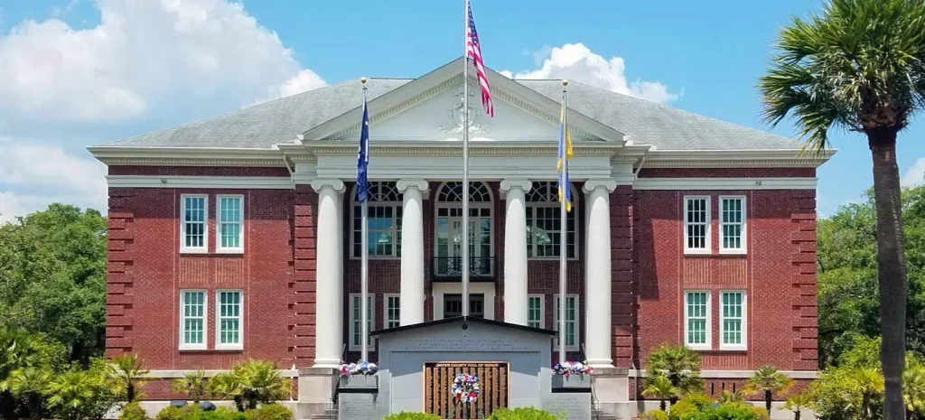 Two-story red-brick Jasper County Courthouse with white classical columns, a gray pediment roof, and an American flag plus two other flags flying in front; a memorial plaque and wreaths stand at the base, with palm trees and landscaping nearby