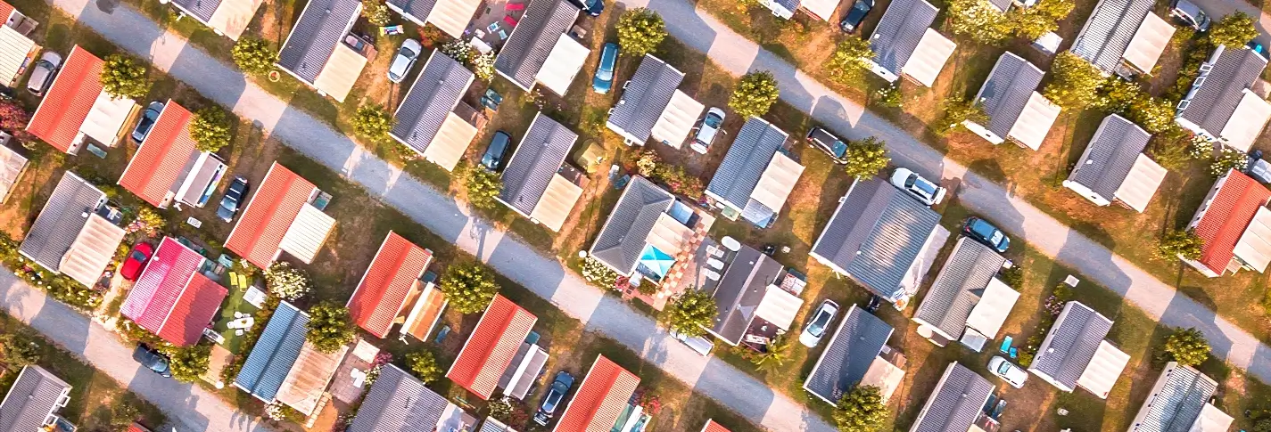 Overhead view of a residential neighborhood with rows of single-story houses featuring red, gray, and tan roofs, parked cars, trees, and paved streets.