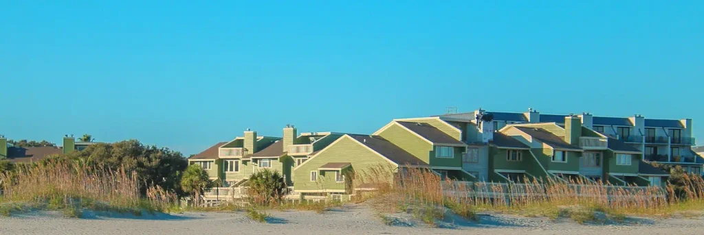 Row of multi-story green coastal houses and condominiums with balconies and chimneys, set behind sandy dunes and tall beach grass under a clear blue sky.