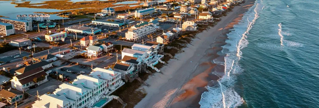 Aerial view of a beachfront community with colorful houses, hotels, and buildings along a sandy shoreline where waves meet the Atlantic Ocean under golden late-day light.