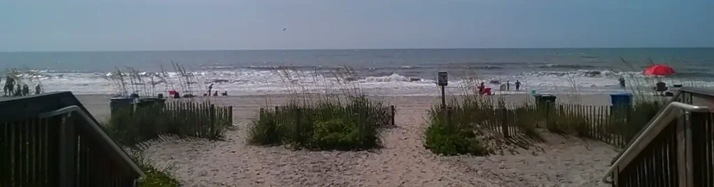 Sandy path through dunes and sea grass leading to a lively beach with people, umbrellas, and ocean waves.  


