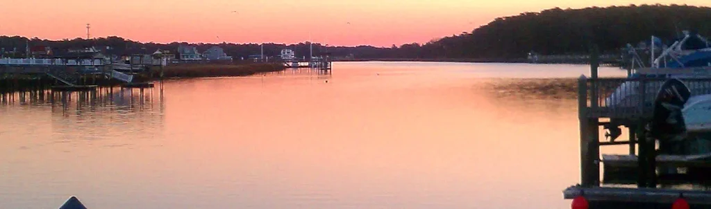 Calm harbor at sunset with pink and orange sky reflecting on still water, wooden docks and boats on the left, houses and trees along the distant shoreline.
