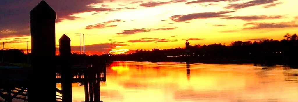 Silhouetted wooden pilings and dock in the foreground against a vivid orange-yellow sunset sky, with the sun near the horizon, its reflection on the water, and a small lighthouse visible in the distance among trees.