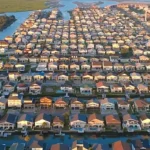Aerial view of a dense coastal neighborhood of houses with varied roofs, interwoven canals and waterways, green marshland on the left, and tall beachfront high-rises on the right near the ocean.