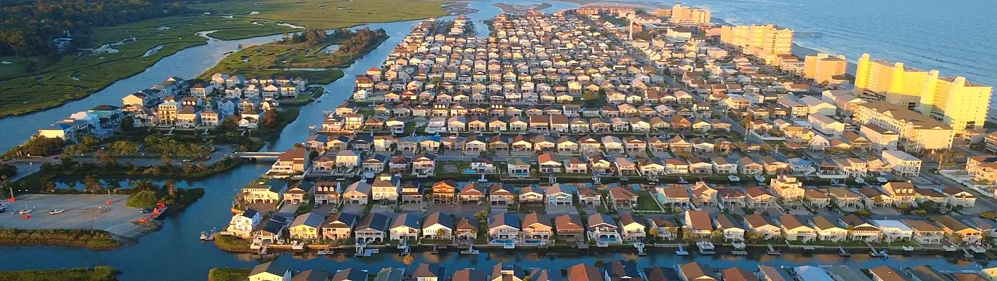 Aerial view of a dense coastal neighborhood of houses with varied roofs, interwoven canals and waterways, green marshland on the left, and tall beachfront high-rises on the right near the ocean.
