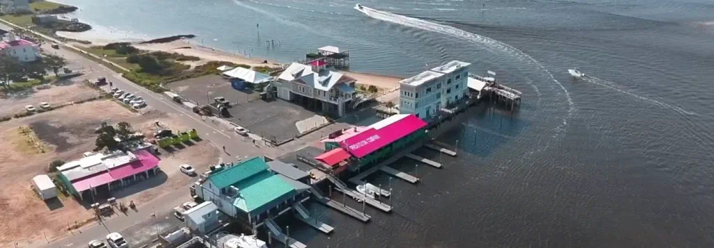 High-angle view of the waterfront showing the green Provision Company building with red roof, docks, boats, surrounding structures, a sandy beach area, and a boat creating a wake on the water.