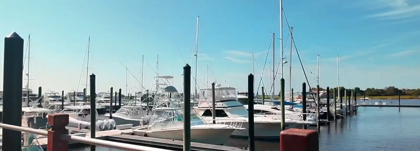 Rows of white boats and sailboats docked at wooden piers with dark pilings under a clear blue sky; calm water and distant shoreline in the background.