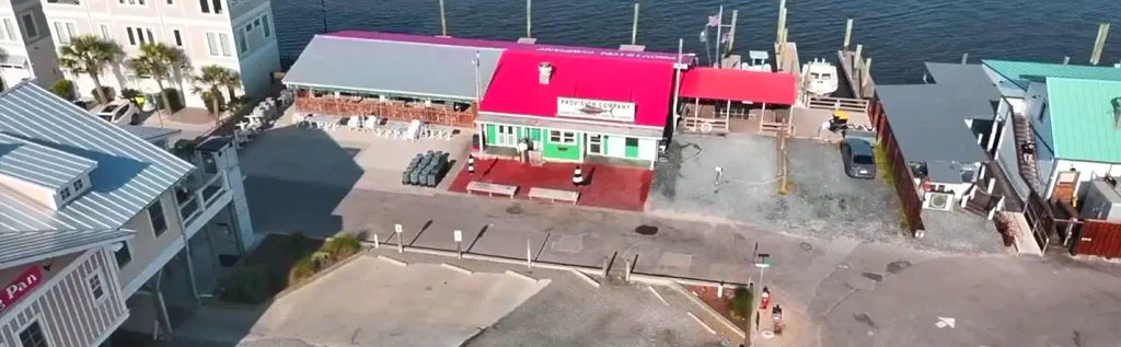 Overhead shot of the green Provision Company building with red roof, docks, and waterfront, surrounded by parking areas, other buildings, and boats along the shoreline.