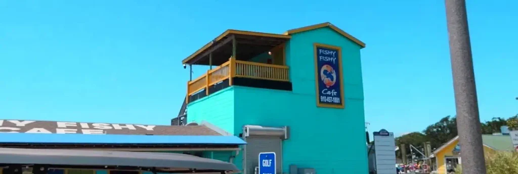 Bright turquoise two-story building with yellow trim and a vertical blue sign reading “FISHY FISHY Cafe 910-457-1881” with a fish logo. Upper deck with wooden railing is visible against a clear blue sky.