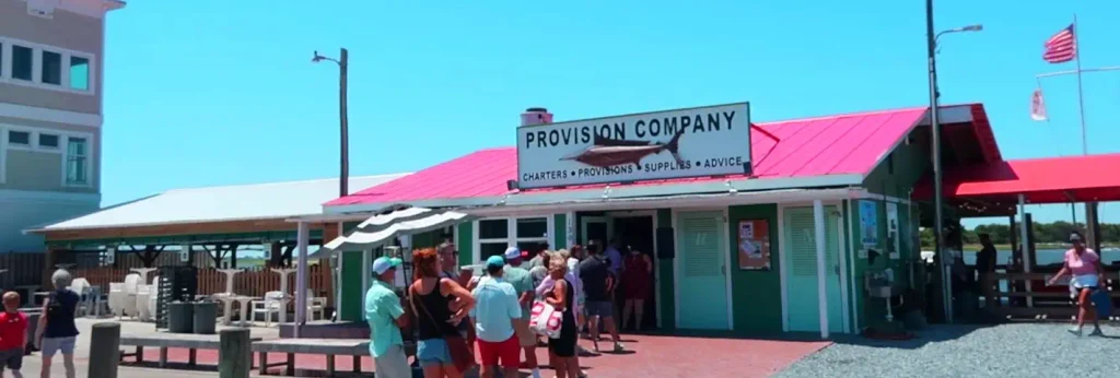 Green building with bright red metal roof and a large sign reading “PROVISION COMPANY – CHARters • Provisions • Supplies • Advice” featuring a fish silhouette. Group of people stands outside the open doorway on a sunny day; outdoor seating and an American flag are visible nearby.