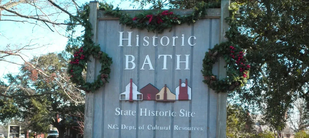 Wooden sign reading “Historic BATH State Historic Site” with illustrations of historic buildings, decorated with green garlands and red berries, under bare tree branches.

