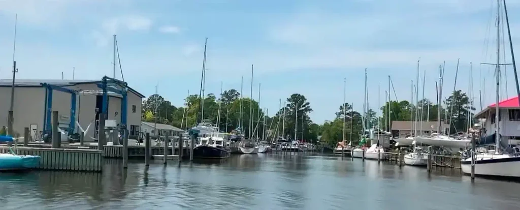 View down a calm marina channel lined with sailboats and powerboats docked on both sides, a large beige building with blue accents on the left, green trees in the background, under a partly cloudy blue sky.

