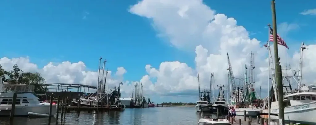 Sunny harbor scene with commercial fishing boats docked on both sides, an American flag flying on the right, a few people standing on the dock, and large white clouds in a bright blue sky.

