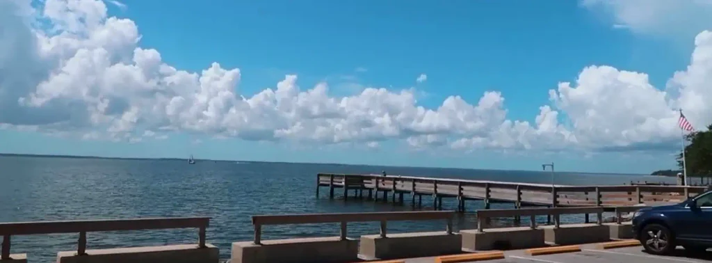 Wide view of a wooden pier extending into calm blue water under a bright sky with puffy white clouds, with a dark car parked near a concrete railing in the foreground.


