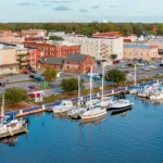 Elevated view of a small waterfront town with brick and colorful buildings, parking lots, a red-roofed pavilion, and numerous boats docked along a long pier on the right.