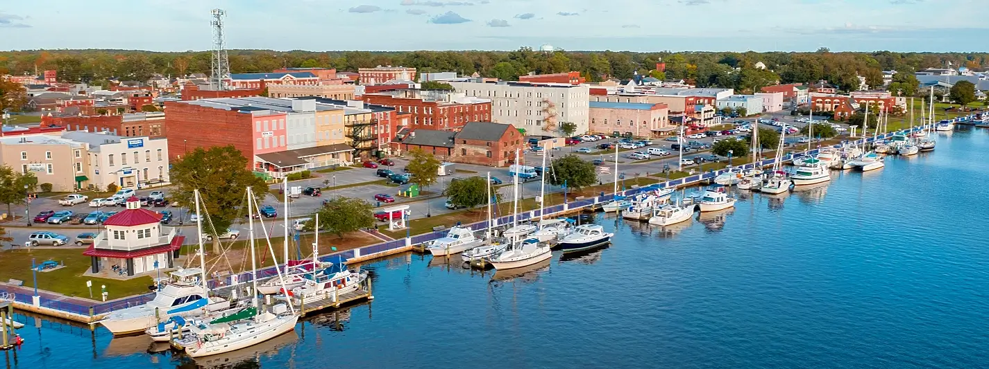 Elevated view of a small waterfront town with brick and colorful buildings, parking lots, a red-roofed pavilion, and numerous boats docked along a long pier on the right.