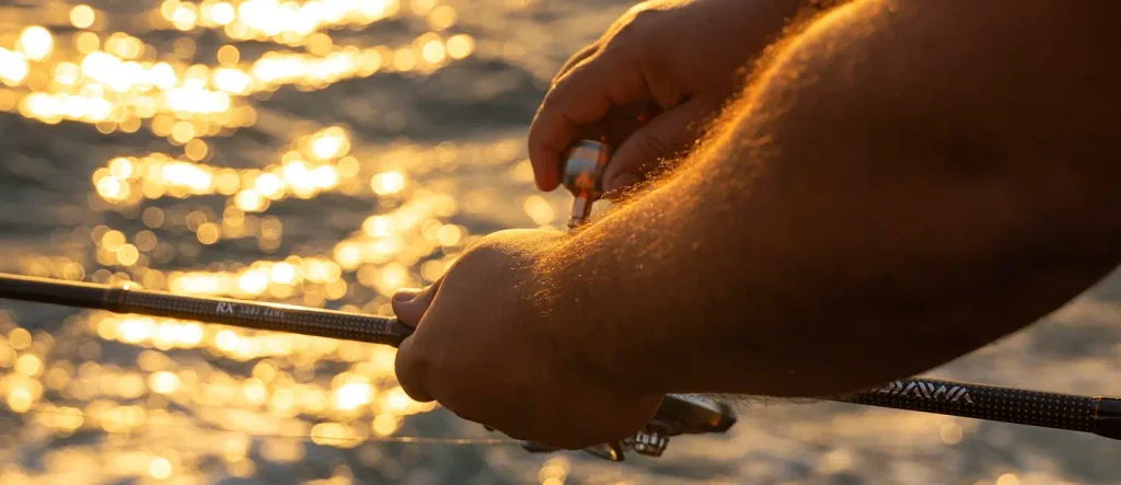Close-up of a person’s hands gripping a fishing rod and spinning reel in warm golden light, with sparkling bokeh reflections on the water in the background.

