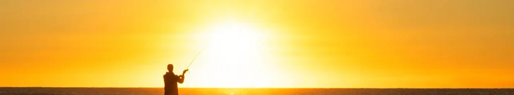 Silhouette of an angler holding a fishing rod against a brilliant yellow-orange sun rising or setting over the ocean horizon.

