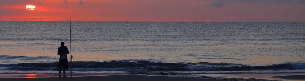 Silhouette of a fisherman standing on the beach at sunset next to a vertical fishing rod, with the sun low on the horizon over calm ocean water reflecting orange and purple light.

