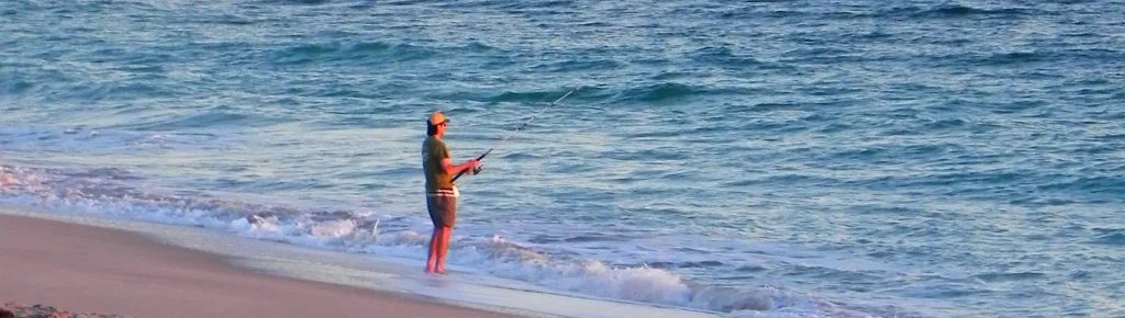 Angler in a green shirt, shorts, and cap standing at the water’s edge on a sandy beach, holding a fishing rod over calm turquoise ocean waves.

