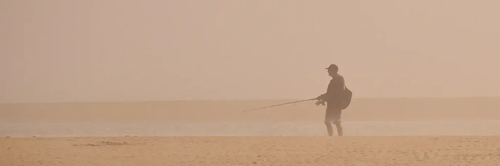 Person with a backpack and fishing rod walking along a wide sandy beach under thick, hazy fog that obscures the horizon and distant water.


