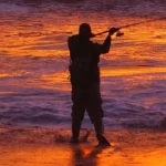 Silhouette of a fisherman standing in shallow surf at sunset, holding a fishing rod extended over orange-glowing waves and wet sand.
