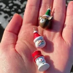 A person’s open hand holds three small painted Christmas ornaments against a background of dark wet sand and white ocean foam: a brown bear in a green-and-white hat, a Santa Claus face with red hat, and a snowman with a red top hat.